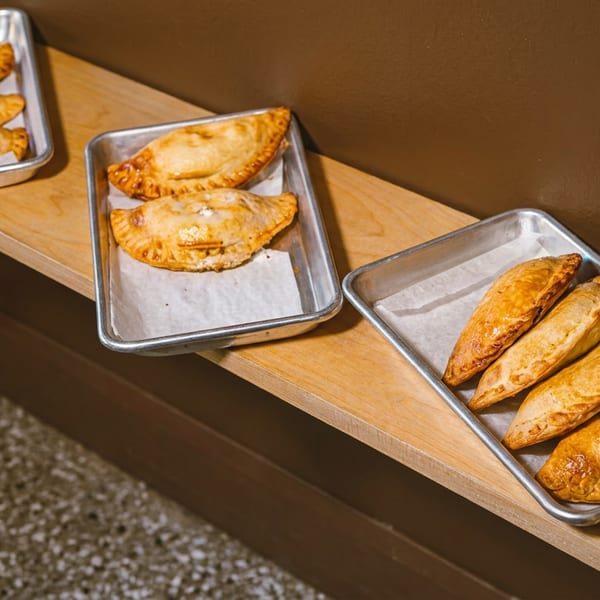 Golden-brown empanadas in metal trays on a wooden shelf, with a dark brown wall behind them.